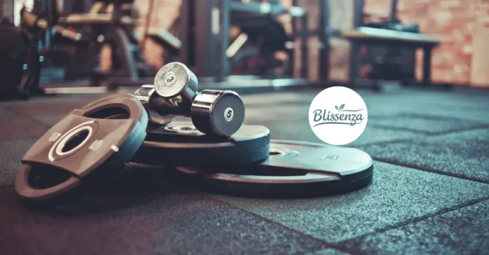 A close-up of gym equipment featuring iron weight plates and two chrome dumbbells on a rubber gym floor.