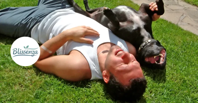 A man lying on green grass in the sun with a dog lying on its back next to him, both looking relaxed and happy.